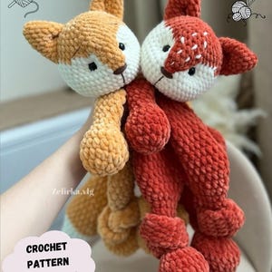 May include: Two crocheted fox plushies, one orange and one red, with white faces and black eyes. The plushies are made with a fuzzy yarn and have a textured appearance. The plushies are being held by a hand.