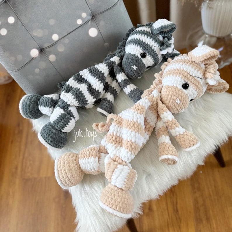 May include: Two handmade crochet animal toys: a gray and white striped zebra and a tan and white striped giraffe. The toys are lying on a white furry surface. The text "juk.toys" is visible on the lower left.