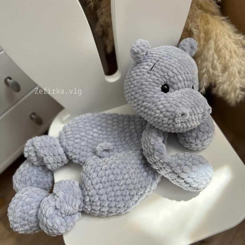 Puede incluir: Un juguete de peluche de hipop&oacute;tamo gris tejido a crochet con una superficie texturizada. El juguete est&aacute; acostado en una silla blanca.