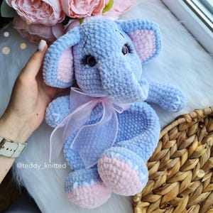 May include: A crocheted blue elephant plush toy with pink ears and feet. The elephant is wearing a pink ribbon around its neck. The toy is sitting on a woven basket.