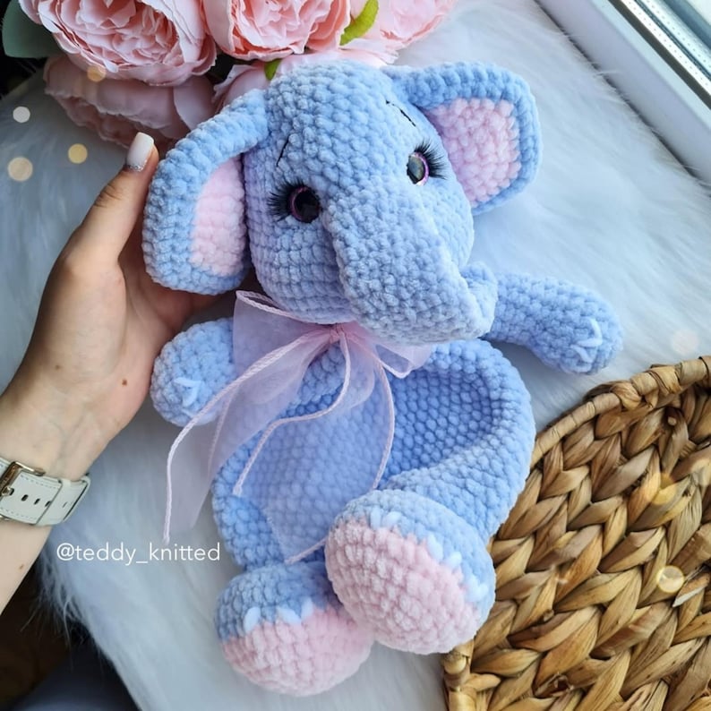 May include: A crocheted blue elephant plush toy with pink ears and feet. The elephant is wearing a pink ribbon around its neck. The toy is sitting on a woven basket.
