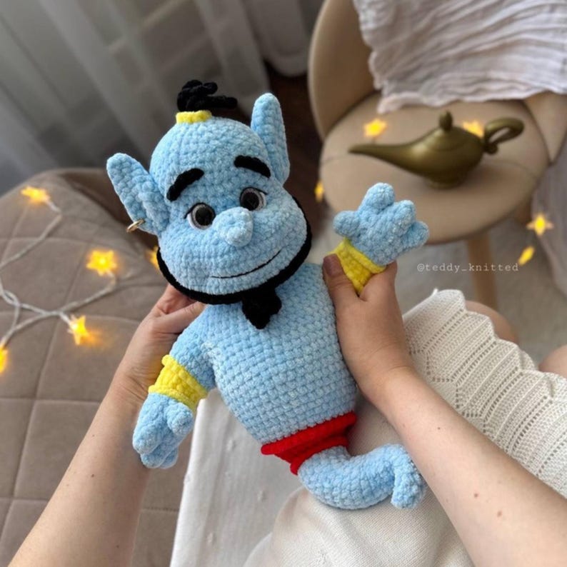 May include: A light blue crocheted plush doll featuring a black beard, black eyebrows, and a small black topknot. The doll has yellow wrist cuffs and a red waistband. The plush has large eyes with black pupils and a soft, textured yarn finish.