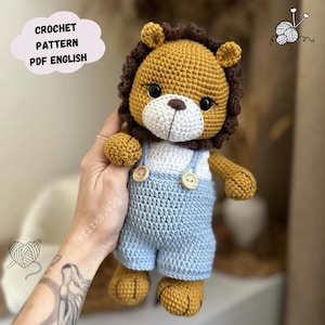 May include: Crochet pattern for a lion amigurumi toy wearing blue dungarees. The lion has a brown mane, white face, and black eyes. The pattern is available in PDF format and is written in English.
