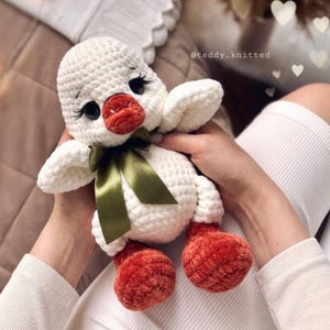 May include: A handmade white crochet duckling with a red beak and feet, black eyes, and long eyelashes. The duckling has a green satin ribbon tied in a bow around its neck. The image is of a soft toy.