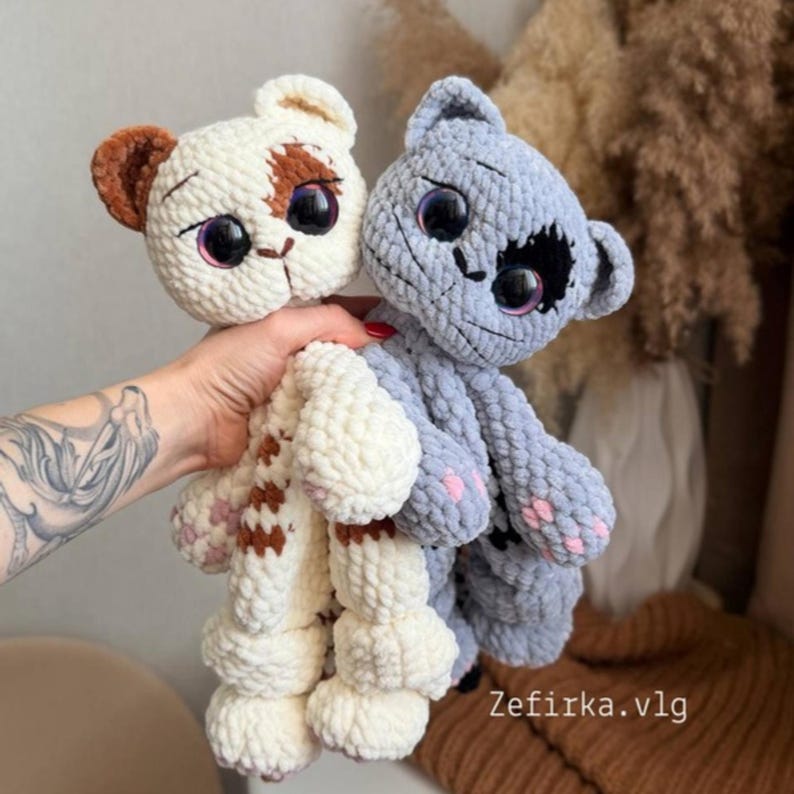 May include: Two handmade crochet cat plushies. One is white with brown accents, the other is gray. Both have large, expressive eyes and pink paw pads. The plushies are soft and cuddly, perfect for a child's toy or a decorative item.