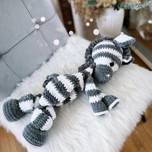May include: A handmade plush zebra toy in gray and white stripes, lying on a white fluffy surface. The zebra has a gray mane, ears, and hooves. The text "juk.toys" is visible on the image.