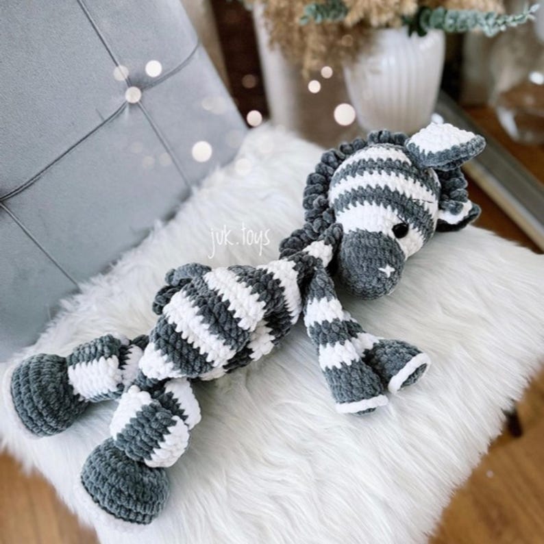 May include: A handmade plush zebra toy in gray and white stripes, lying on a white fluffy surface. The zebra has a gray mane, ears, and hooves. The text "juk.toys" is visible on the image.