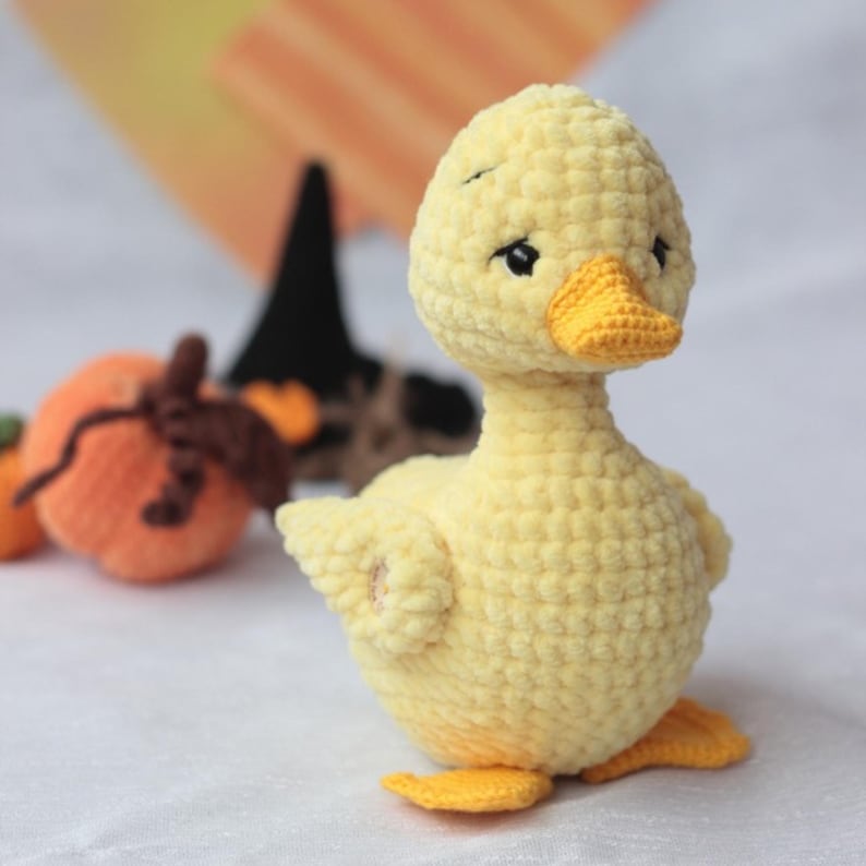 Peut inclure: Un caneton jaune en peluche fait &agrave; la main, avec un bec et des pieds jaune vif. Le caneton a de petits yeux noirs et est fait d'un fil doux et textur&eacute;. L'arri&egrave;re-plan comprend une petite citrouille orange et un chapeau de sorci&egrave;re noir.