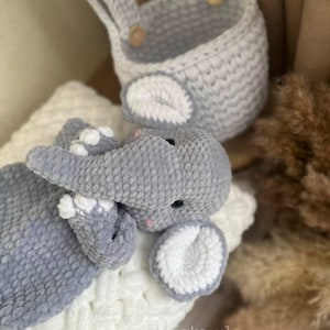 May include: A handmade gray crochet elephant toy with white accents, lying on a white textured surface. A matching gray crochet basket with wooden buttons is in the background. The elephant has black eyes and pink details.