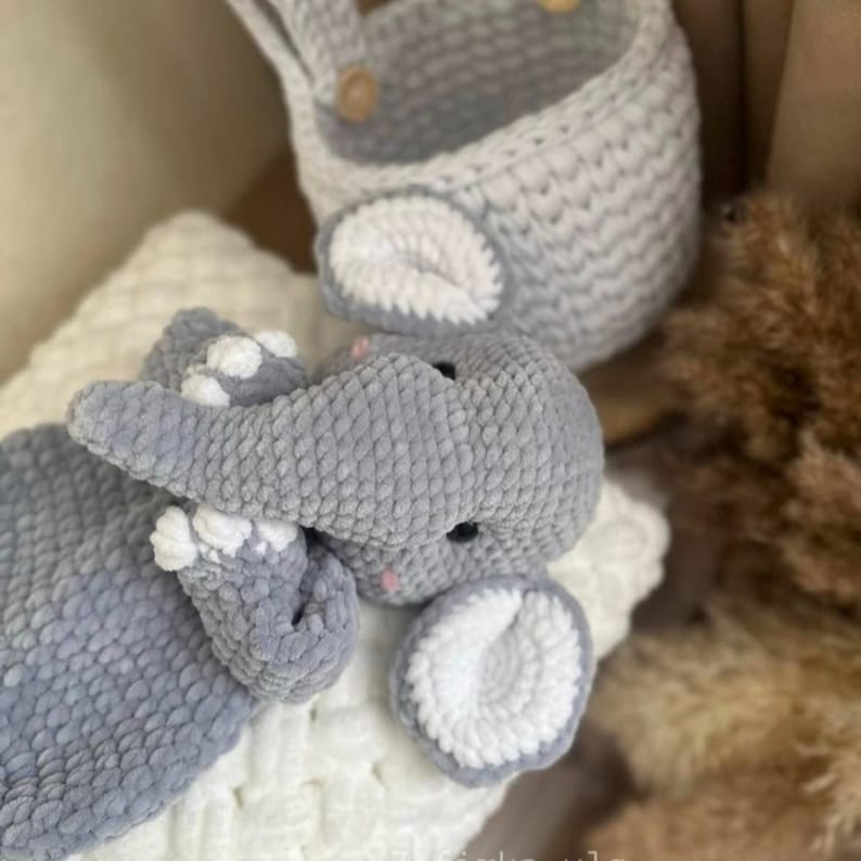 May include: A handmade gray crochet elephant toy with white accents, lying on a white textured surface. A matching gray crochet basket with wooden buttons is in the background. The elephant has black eyes and pink details.