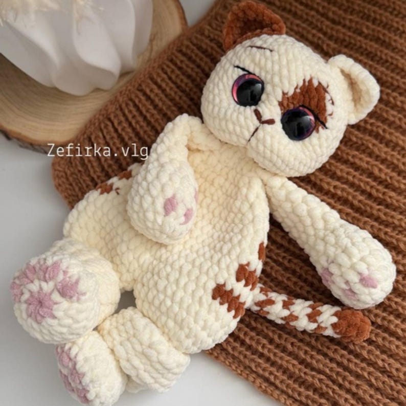 May include: A handmade, cream-colored crochet cat with brown spots and accents. The cat has large, expressive eyes and pink paw pads. The text "Zefirka.vlg" is visible on the left side. The cat is lying on a brown, textured surface.