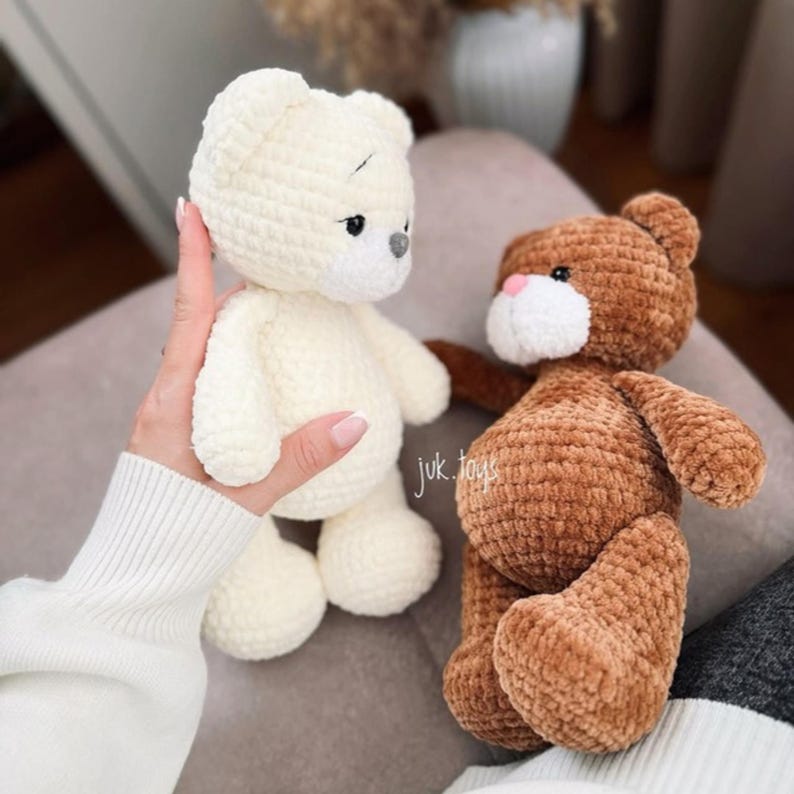 Op de afbeelding: Twee gehaakte teddyberen, een witte en een bruine, zitten op een lichtgrijze ondergrond. De witte beer wordt vastgehouden door een hand. De bruine beer heeft een witte neus en mond.