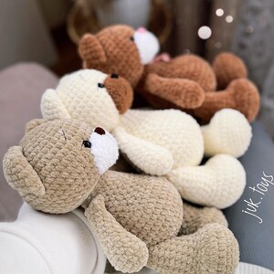 Op de afbeelding: Drie gehaakte teddyberen in verschillende tinten bruin, beige en wit. De beren liggen op een wit oppervlak. De beren hebben zwarte ogen en bruine neuzen.