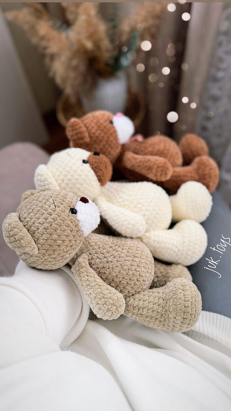 Op de afbeelding: Drie gehaakte teddyberen in verschillende tinten bruin, beige en wit. De beren liggen op een wit oppervlak. De beren hebben zwarte ogen en bruine neuzen.