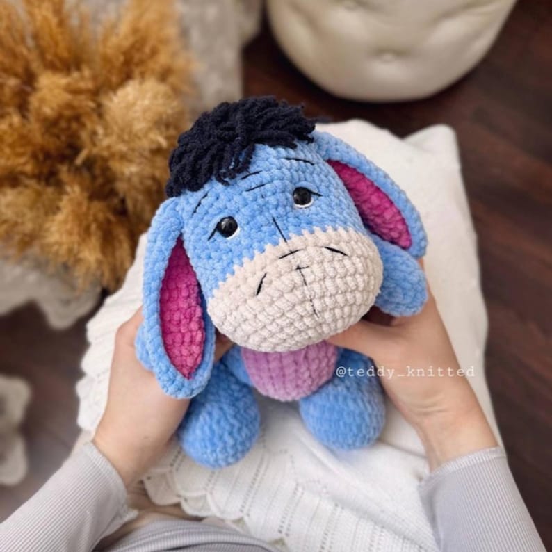May include: A blue crocheted stuffed animal donkey with pink ears, a white face, and black hair. The donkey is being held by a person's hands.