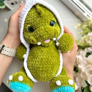 May include: Green crocheted dinosaur plush toy wearing a white crocheted hooded hat with blue accents on the feet. The toy is being held by a person's hands. @teddy_knitted