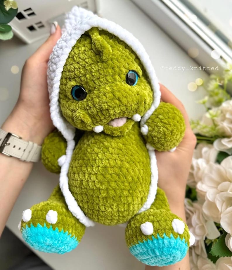 May include: Green crocheted dinosaur plush toy wearing a white crocheted hooded hat with blue accents on the feet. The toy is being held by a person's hands. @teddy_knitted