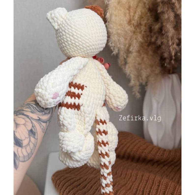 May include: A handmade, cream-colored crochet cat doll with brown striped accents on its body and tail. The cat has pink paw pads and a brown patch on its head. The doll is soft and plush, with a textured appearance.