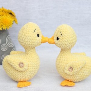 Peut inclure: Deux canetons au crochet jaunes faits &agrave; la main, avec des becs et des pieds oranges, se touchent le bec. Chaque caneton a un bouton sur le c&ocirc;t&eacute; et des yeux noirs. Un vase gris avec des fleurs jaunes est en arri&egrave;re-plan.