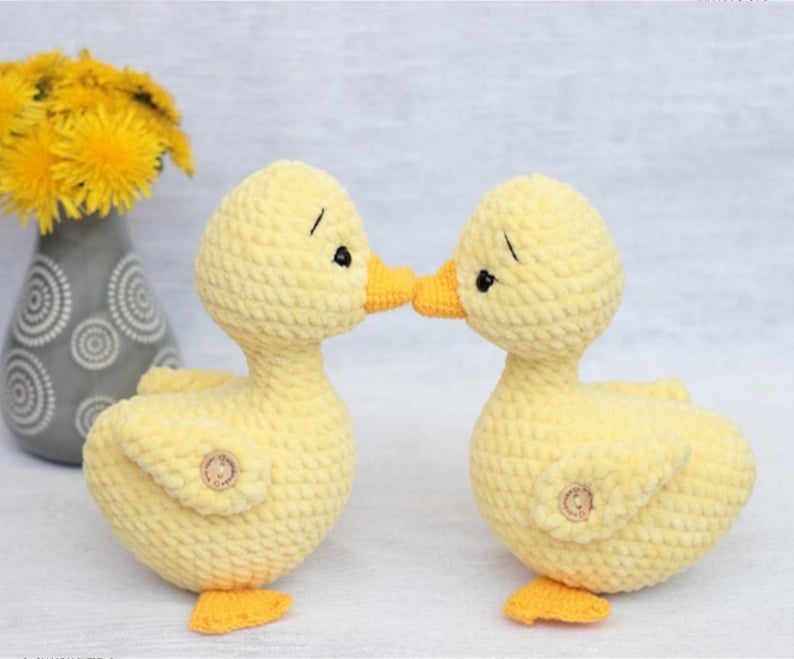 Peut inclure: Deux canetons au crochet jaunes faits &agrave; la main, avec des becs et des pieds oranges, se touchent le bec. Chaque caneton a un bouton sur le c&ocirc;t&eacute; et des yeux noirs. Un vase gris avec des fleurs jaunes est en arri&egrave;re-plan.