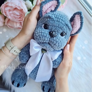 May include: A crocheted plush toy in the shape of a French bulldog. The toy is blue with pink ears and a white bow. The toy is being held by a person's hands.