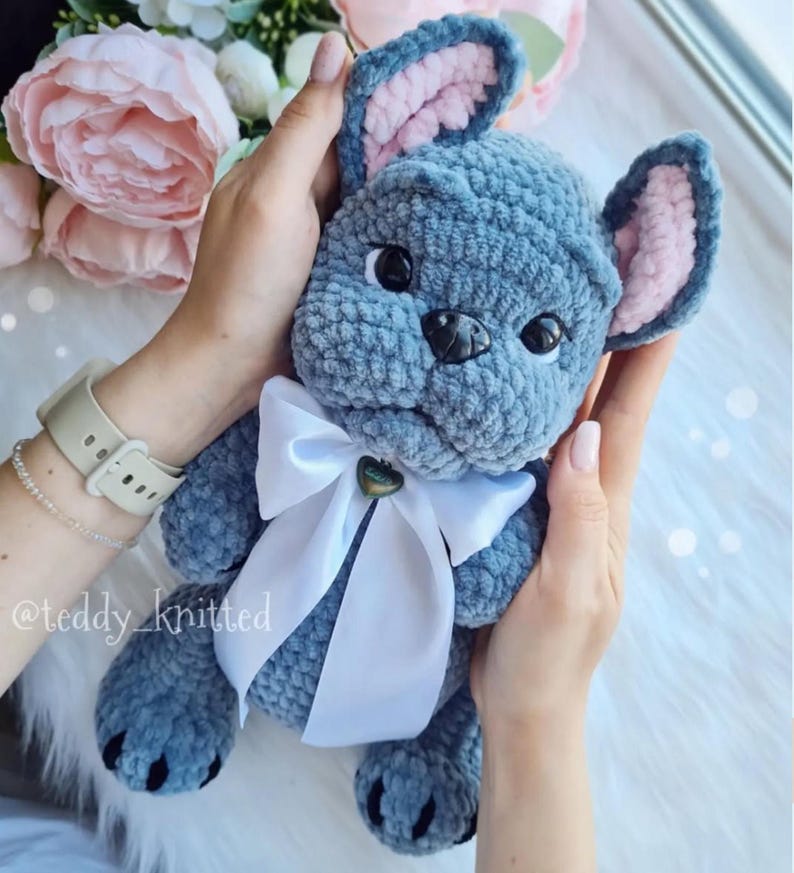 May include: A crocheted plush toy in the shape of a French bulldog. The toy is blue with pink ears and a white bow. The toy is being held by a person's hands.