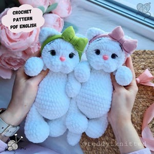 Puede incluir: Dos peluches de gato de ganchillo blanco con lazos verdes y rosas. Los gatos tienen ojos negros y narices rosas. La imagen incluye el texto "CROCHET PATTERN PDF ENGLISH". Los peluches est&aacute;n hechos a mano y parecen suaves.