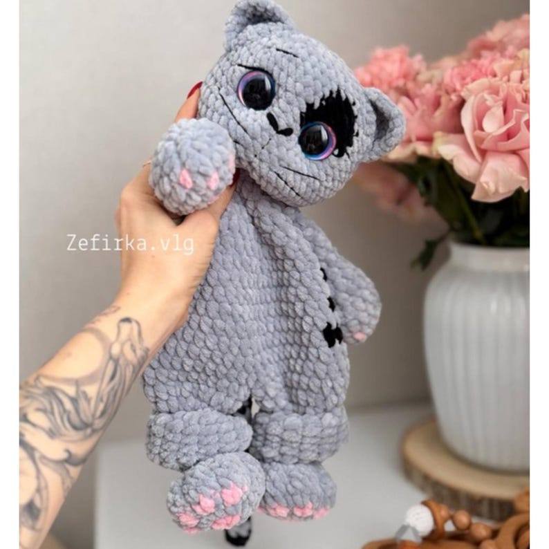 May include: A handmade gray crochet cat plushie with large, iridescent eyes and pink paw pads. The cat has a stitched face and black accents. The plushie is held up by a hand. The text "Zefirka.vlg" is visible.