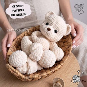 Pode incluir: Um urso de peluche de croché cor de creme com olhos escuros e nariz castanho, repousando dentro de um cesto oval entrançado. A imagem inclui o texto CROCHET PATTERN PDF ENGLISH e Zefirka.vlg, juntamente com ícones de novelos de lã.