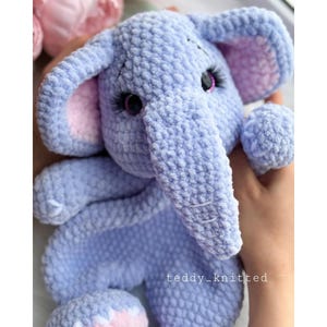 May include: A crocheted plush elephant toy with a light blue body, pink ears, and large purple eyes. The elephant is holding its trunk up and has a textured, fuzzy surface.