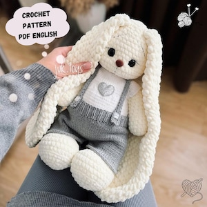 Puede incluir: Un juguete de conejo de crochet en crema y gris. El conejo lleva un peto gris con tirantes y una camisa blanca con un corazón. La imagen incluye el texto "CROCHET PATTERN PDF ENGLISH".