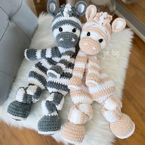 May include: Two handmade crochet zebra plush toys. One is gray and white striped, the other is peach and white striped. Both have black eyes and are sitting on a white fluffy surface. The text "juk.toys" is visible.
