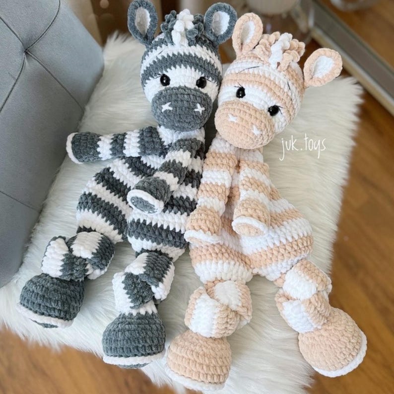 May include: Two handmade crochet zebra plush toys. One is gray and white striped, the other is peach and white striped. Both have black eyes and are sitting on a white fluffy surface. The text "juk.toys" is visible.