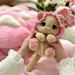 May include: A handmade crochet cat doll in beige with pink accents. The cat has a pink bonnet, pink paws, and a pink nose. The doll is held against a pink background, suggesting a soft and cuddly toy.