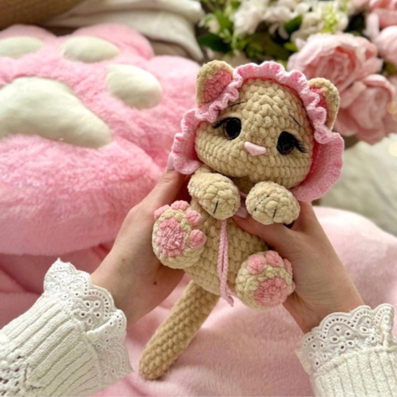 May include: A handmade crochet cat doll in beige with pink accents. The cat has a pink bonnet, pink paws, and a pink nose. The doll is held against a pink background, suggesting a soft and cuddly toy.