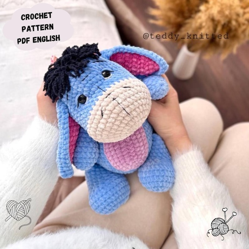 May include: A crocheted blue Eeyore plush toy with pink ears and a black mane. The toy is being held by a person wearing a white sweater. The image includes the text "CROCHET PATTERN PDF ENGLISH" and "@teddy_knitted".