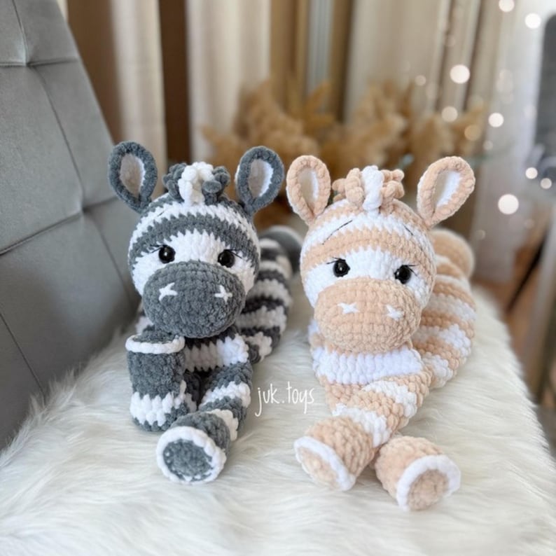 May include: Two plush zebra toys. One is gray and white, the other is peach and white. Both have black eyes and are lying on a white surface. The text "juk.toys" is visible on the right toy.
