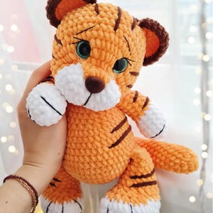 May include: A handmade orange and brown crochet tiger plush toy with black stripes. The tiger has white accents on its paws and muzzle, and green eyes. The ears are brown with orange centers. The tiger is a soft, cuddly toy.