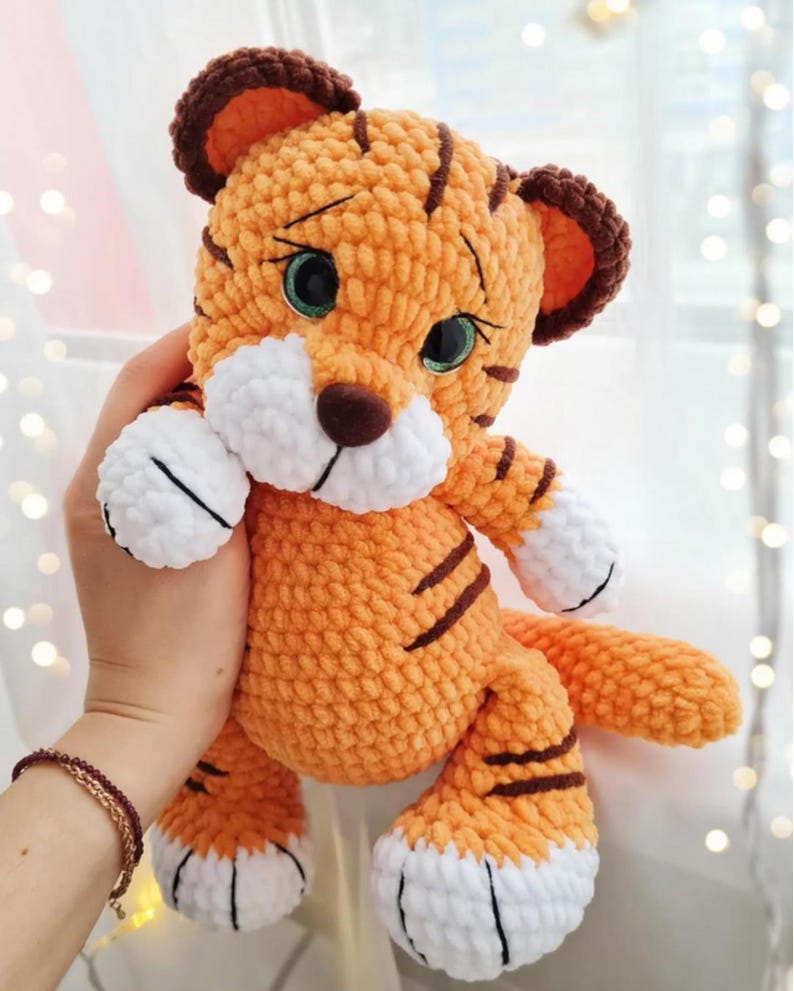 May include: A handmade orange and brown crochet tiger plush toy with black stripes. The tiger has white accents on its paws and muzzle, and green eyes. The ears are brown with orange centers. The tiger is a soft, cuddly toy.