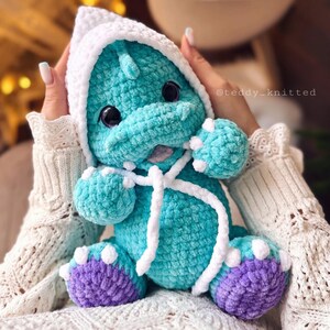 May include: A teal blue crocheted dinosaur plush toy wearing a white crocheted hooded hat with purple feet. The toy is being held by a person's hands.