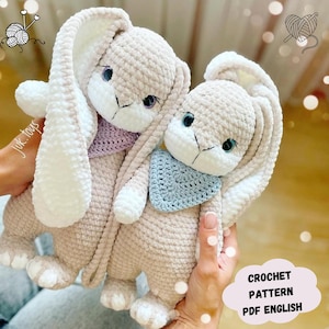 Op de afbeelding: Twee gehaakte konijnen knuffels met witte vacht accenten op hun oren en poten. De konijnen dragen gehaakte slabben, een in lichtpaars en de andere in lichtblauw. De afbeelding bevat de tekst "CROCHET PATTERN PDF ENGLISH".