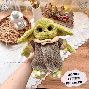 May include: A crocheted doll resembling a character from a popular science fiction series. The doll is primarily green and brown, with large black eyes and a tan collar. The doll is wearing a brown coat. The text "CROCHET PATTERN PDF ENGLISH" is visible.