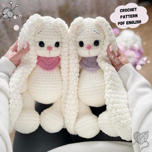 Puede incluir: Dos peluches de conejo de crochet blanco con orejas largas y caídas. Los conejos tienen bufandas rosa y morada y ojos negros. La imagen incluye el texto "CROCHET PATTERN PDF ENGLISH" y "juk.toys".
