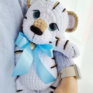 May include: A handmade white tiger plush toy with black stripes, tan ears, and blue eyes. The tiger has a light blue ribbon tied around its neck. A small gold charm is attached to the ribbon. The plush is soft and cuddly.