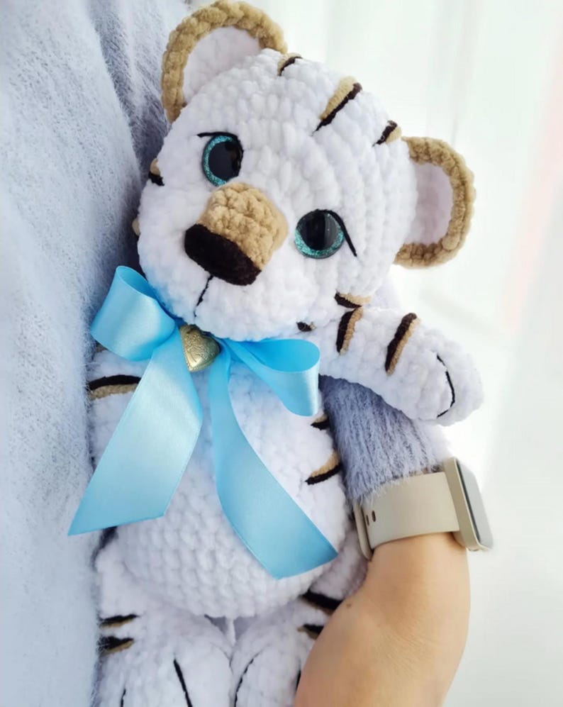 May include: A handmade white tiger plush toy with black stripes, tan ears, and blue eyes. The tiger has a light blue ribbon tied around its neck. A small gold charm is attached to the ribbon. The plush is soft and cuddly.
