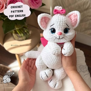 May include: A white crochet cat plush toy with a pink bow on its head and a pink collar. The cat has large blue eyes, long eyelashes, a pink nose, and embroidered whiskers. A text bubble in the corner reads CROCHET PATTERN PDF ENGLISH.