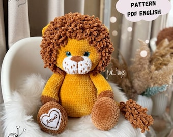 May include: A handmade crochet lion plush toy with a golden yellow body and a brown mane. The lion has blue eyes and a white muzzle. The text "CROCHET PATTERN PDF ENGLISH" is in a cloud-shaped graphic. The lion is sitting on a white fluffy surface.
