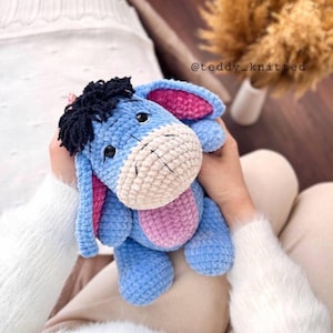 May include: A blue and pink crocheted stuffed animal resembling Eeyore from Winnie the Pooh. The animal has a long face, big ears, and a black mane.