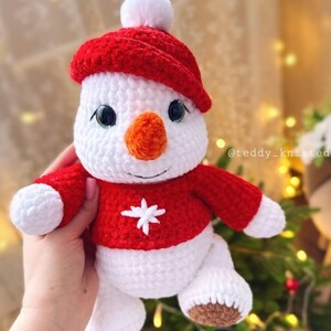 May include: A white crocheted snowman wearing a red crocheted hat and sweater with a white snowflake design. The snowman has a carrot nose and black eyes.