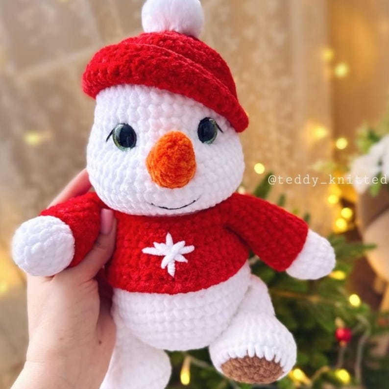 May include: A white crocheted snowman wearing a red crocheted hat and sweater with a white snowflake design. The snowman has a carrot nose and black eyes.